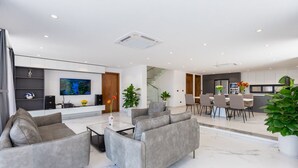 A modern living room with a grey sofa set, a coffee table, and a flat-screen TV. There is a dining area with a table and chairs, and a kitchen area with cabinets and a countertop. - Tran Duy Luxury Villa 3 (Ho Chi Minh City)