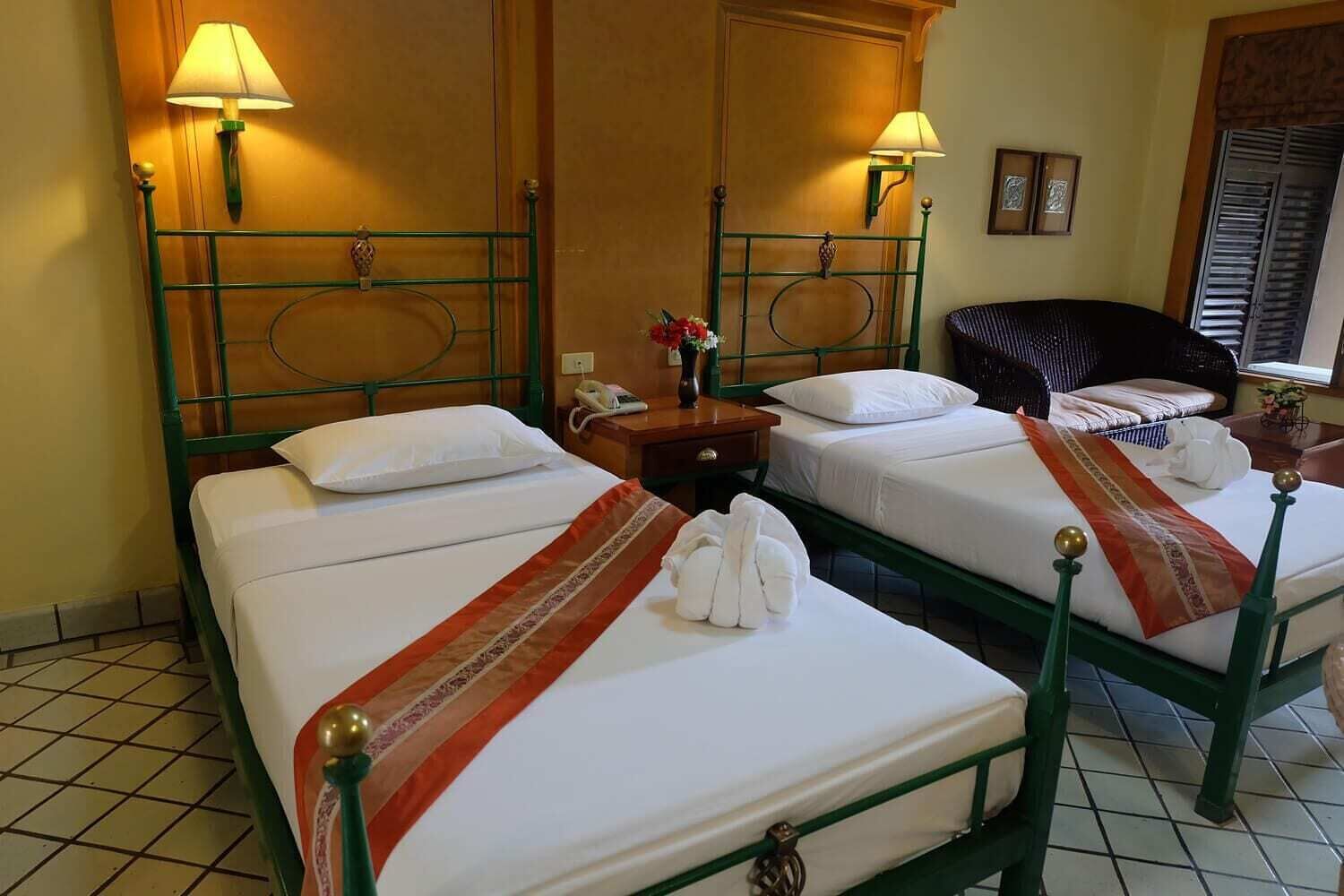 A hotel room with two single beds, each with a green metal frame and white bedding. There are two bedside lamps, a wooden nightstand with a phone, and a small table with a vase of flowers.