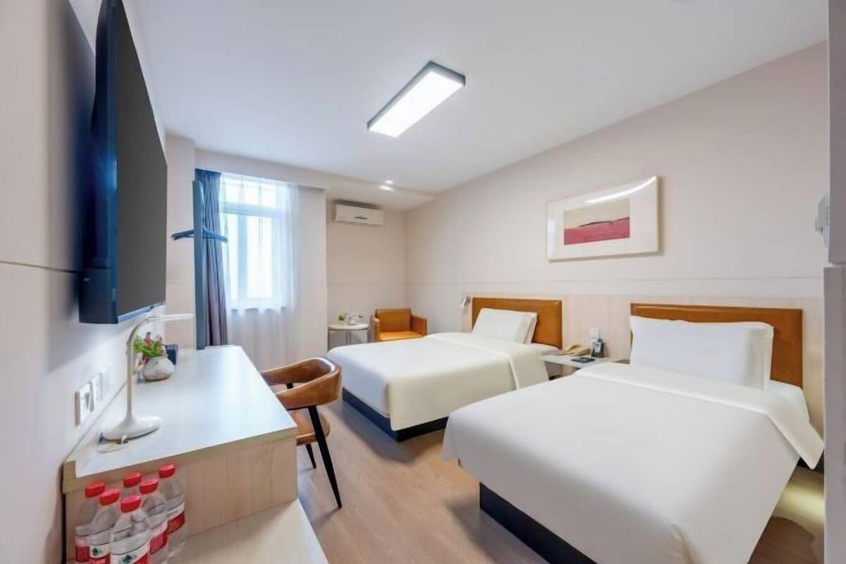 A hotel room with two beds, a desk with a chair, a TV, a framed picture on the wall, and a window with curtains.