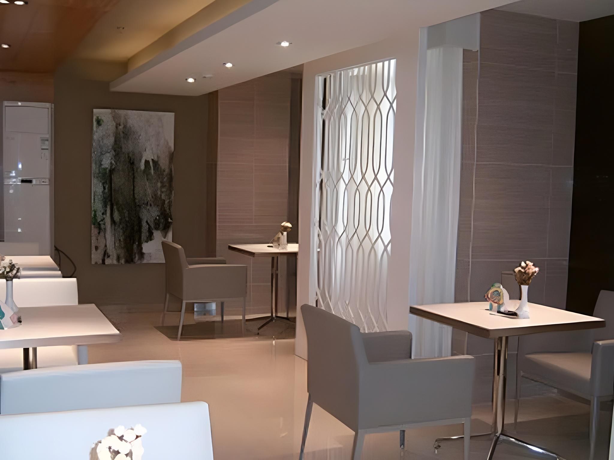 A modern dining area with white tables and chairs, a large abstract painting on the wall, and a decorative window with a geometric pattern.
