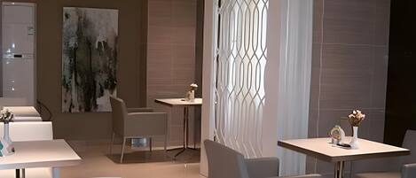 A modern dining area with white tables and chairs, a large abstract painting on the wall, and a decorative window with a geometric pattern.