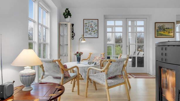 A dining area with wicker chairs, a round table, and a lamp. There is a fireplace and a large window with a view of trees. - Lovely home in ølsted with kitchen (Ølsted)