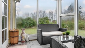 A sunroom with a wicker sofa, a glass table, and a view of a lake surrounded by trees. - Lovely home in ølsted with kitchen (Ølsted)