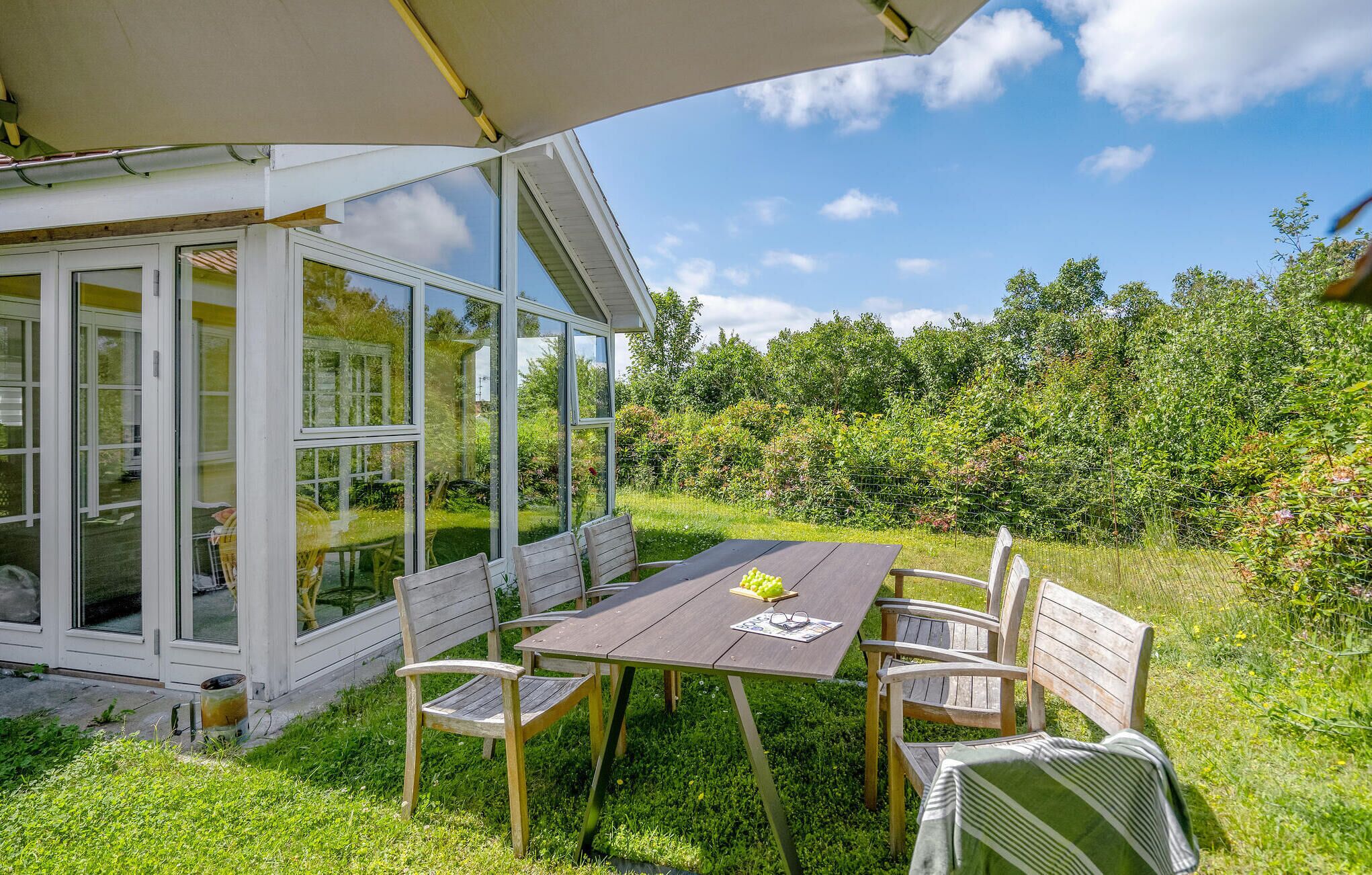 A patio with a table and chairs under a canopy, surrounded by a garden and a white building with glass doors.