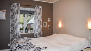 Ein Schlafzimmer mit einem Bett, einem Fenster mit Vorhängen, einem Nachttisch mit Lampe und einer Wand mit gerahmten Bildern.