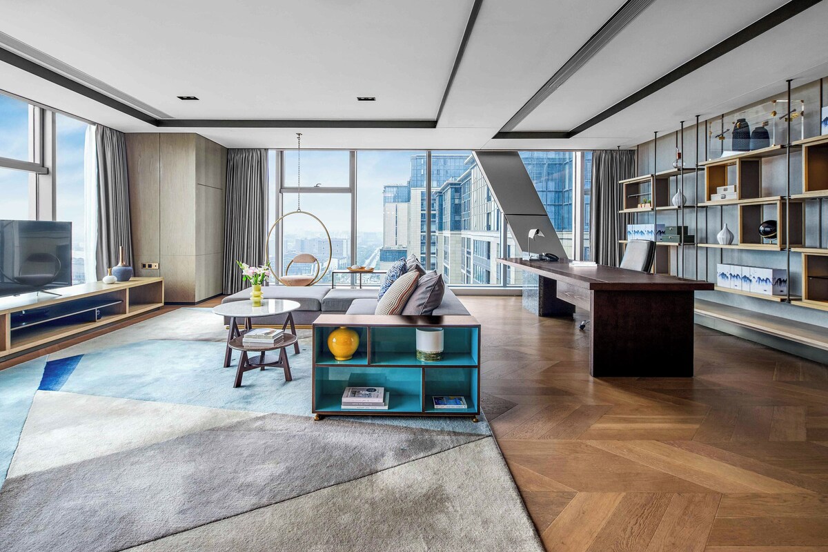 A modern living room with a large window, a sofa, a coffee table, and a bookshelf.