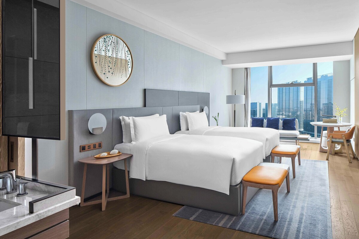 A modern hotel room with two beds, a desk, a chair, and a view of the city skyline.