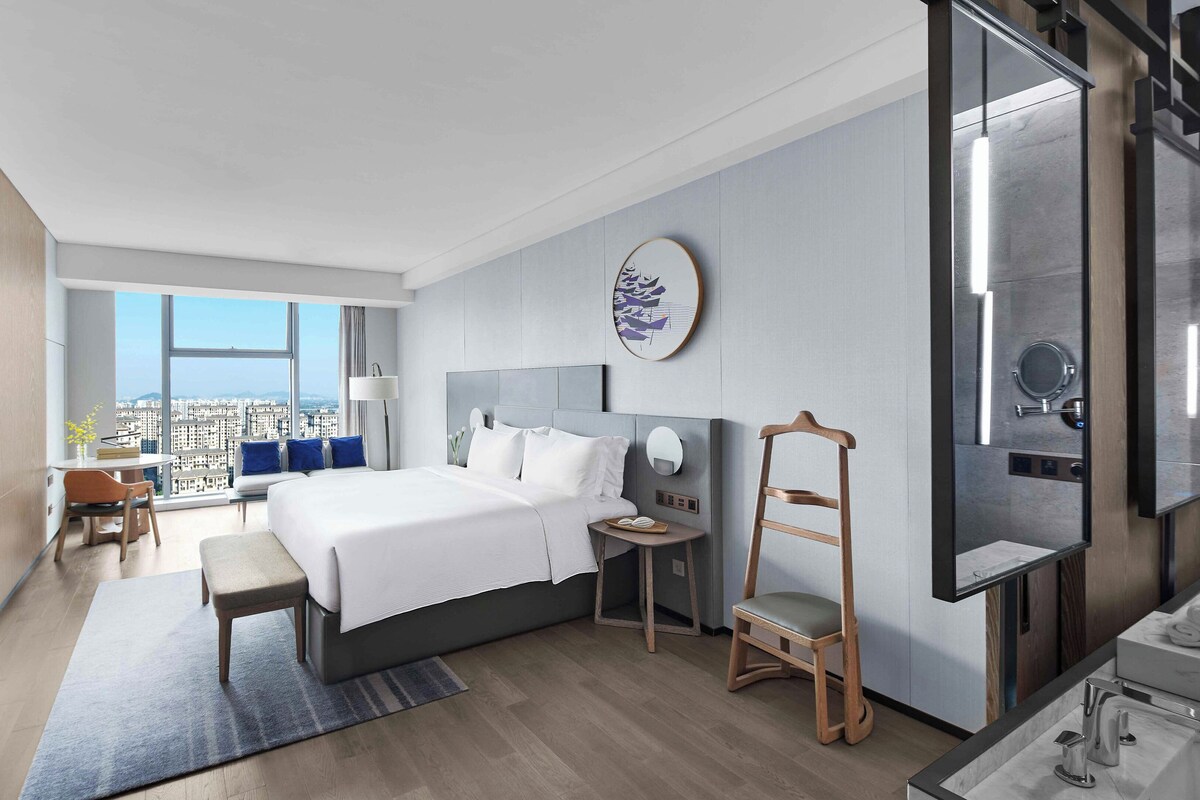 A modern hotel room with a large bed, a desk, a chair, a small table, a mirror, and a view of the city.