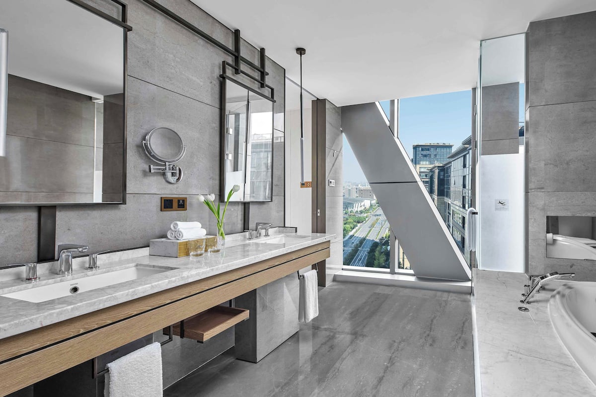 A modern bathroom with a large mirror, two sinks, a bathtub, and a view of the city through a large window.