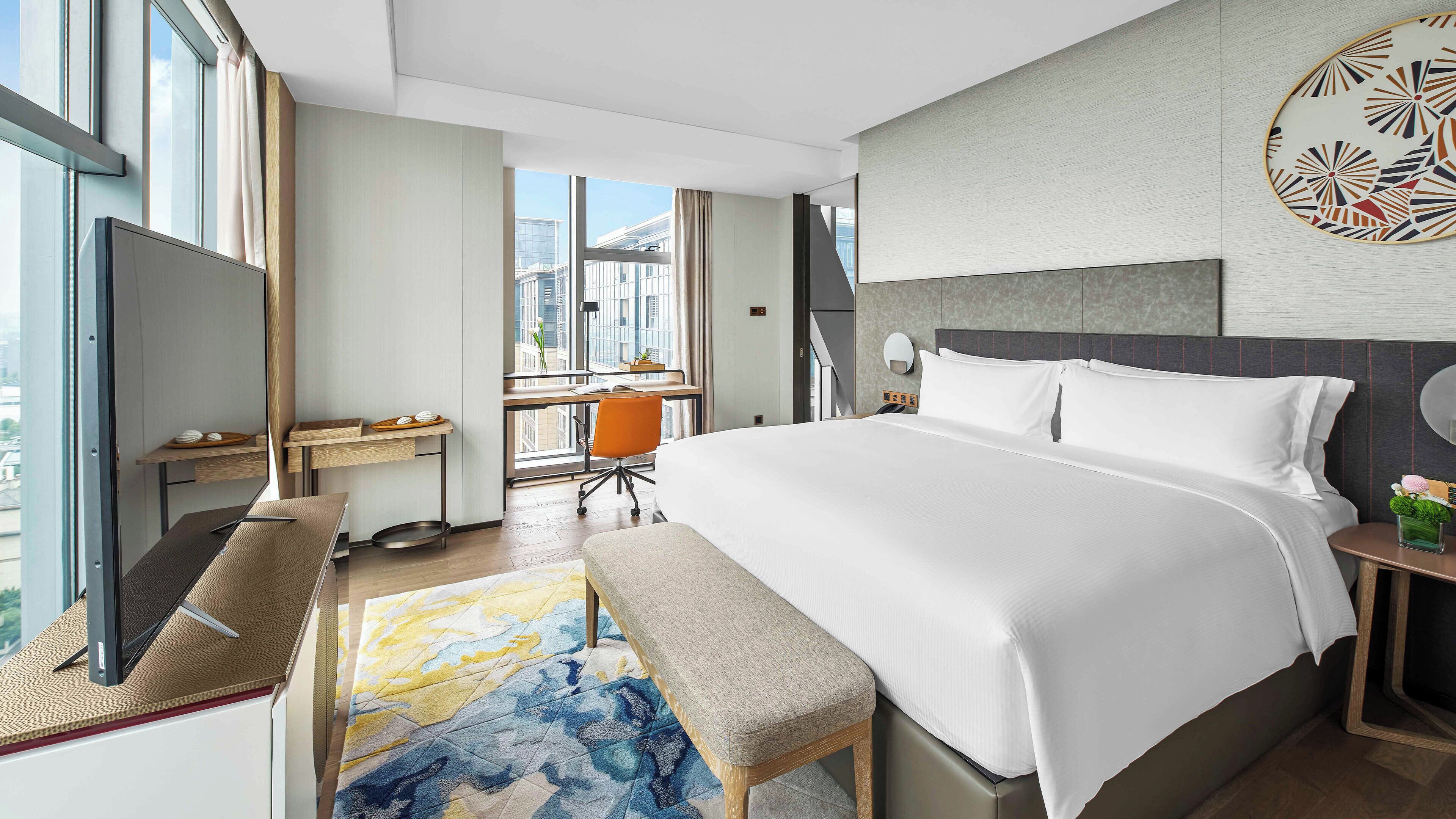 Una habitación de hotel moderna con una cama grande, un escritorio y una silla. Se ve la ciudad a través de la ventana.