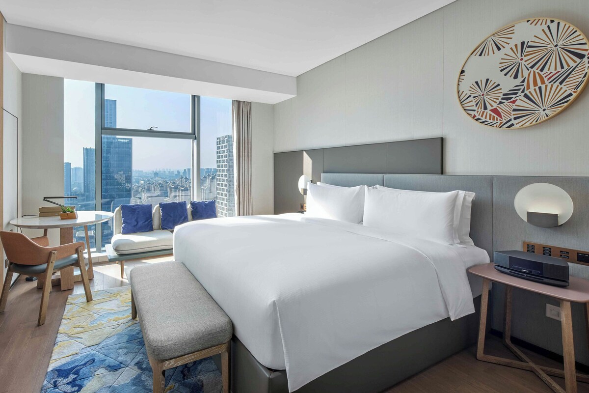 Une chambre d’hôtel moderne dotée d’un grand lit, d’un bureau, d’une chaise, d’une télévision et offrant une vue sur le panorama urbain.