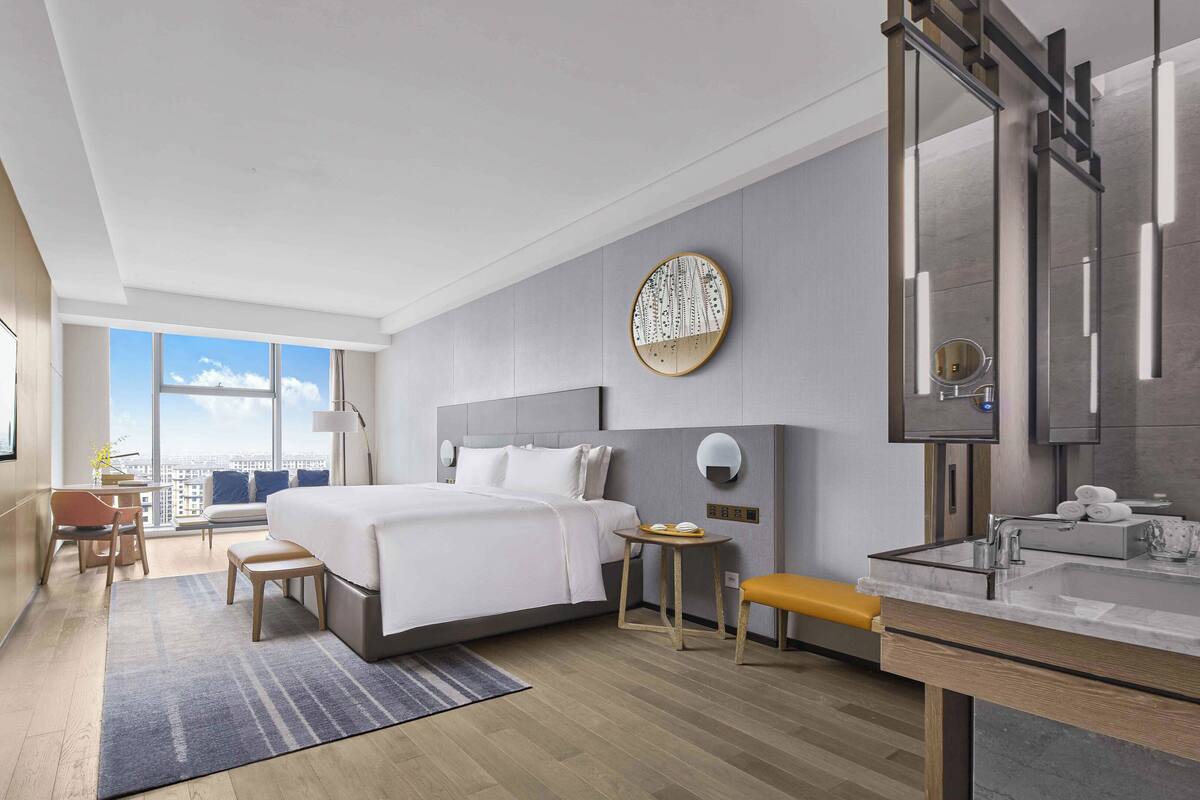 A modern hotel room with a large bed, a desk, a chair, a small table, and a bathroom with a mirror and towel rack.