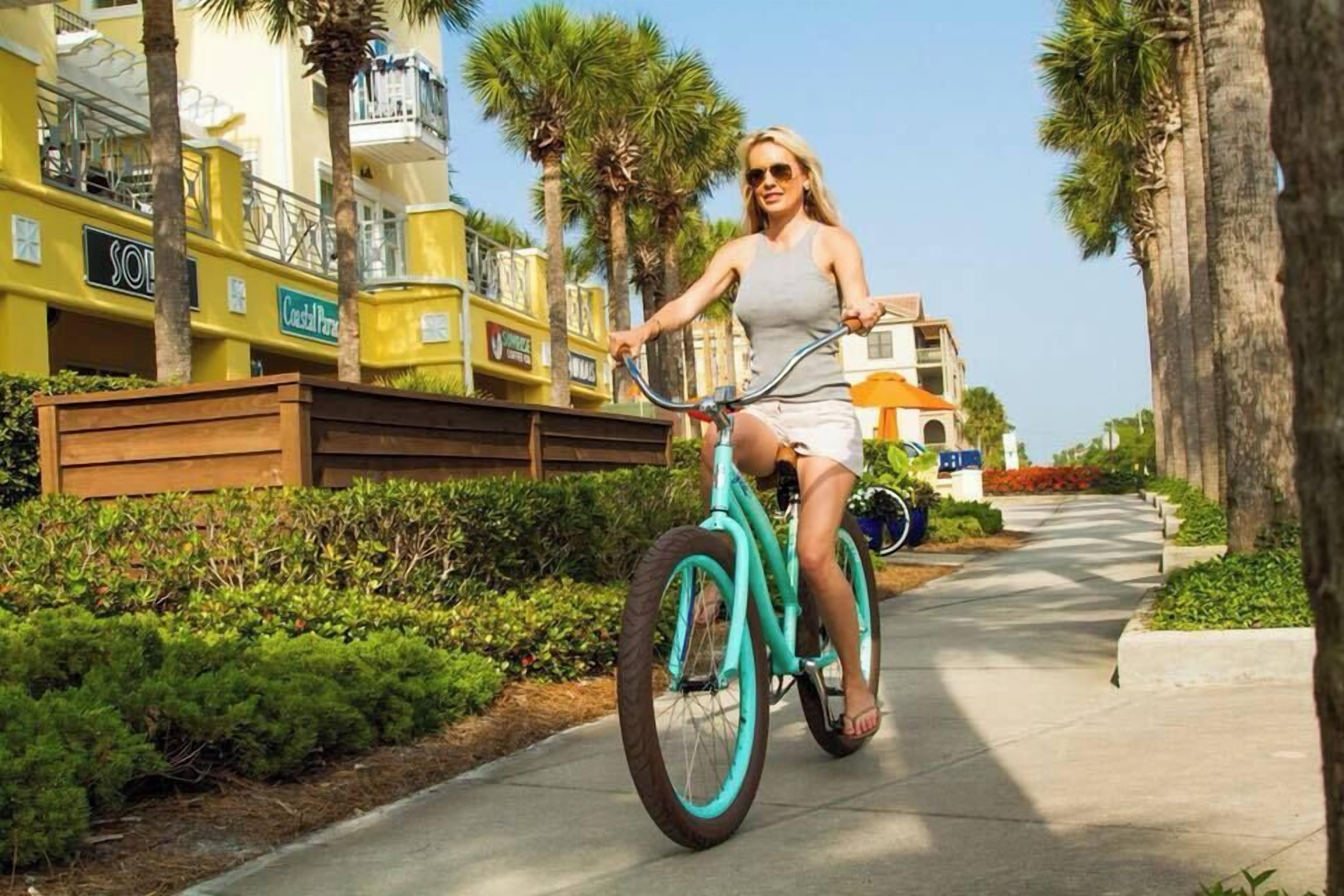 Une personne à vélo, de couleur turquoise, circule sur un trottoir bordé de palmiers et d’immeubles.