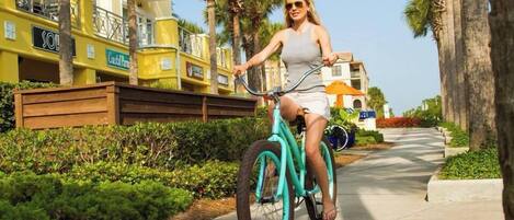Une personne à vélo, de couleur turquoise, circule sur un trottoir bordé de palmiers et d’immeubles.