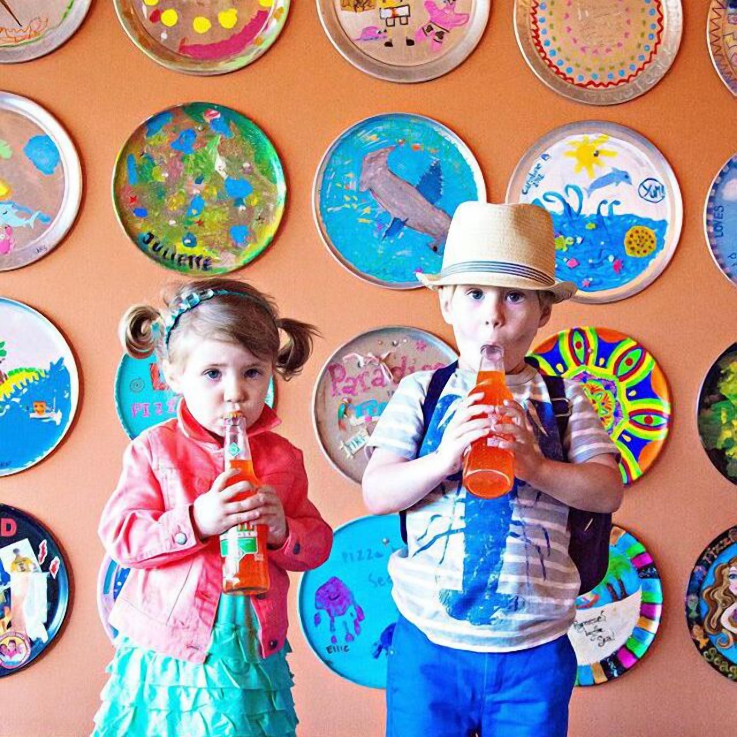 Two children holding bottles, with colorful plates on the wall behind them.