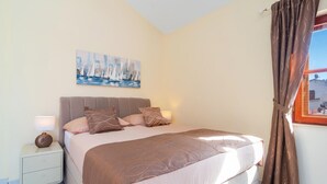 Une chambre à coucher avec un lit, une table de chevet, une lampe, un tableau et une fenêtre avec des rideaux.