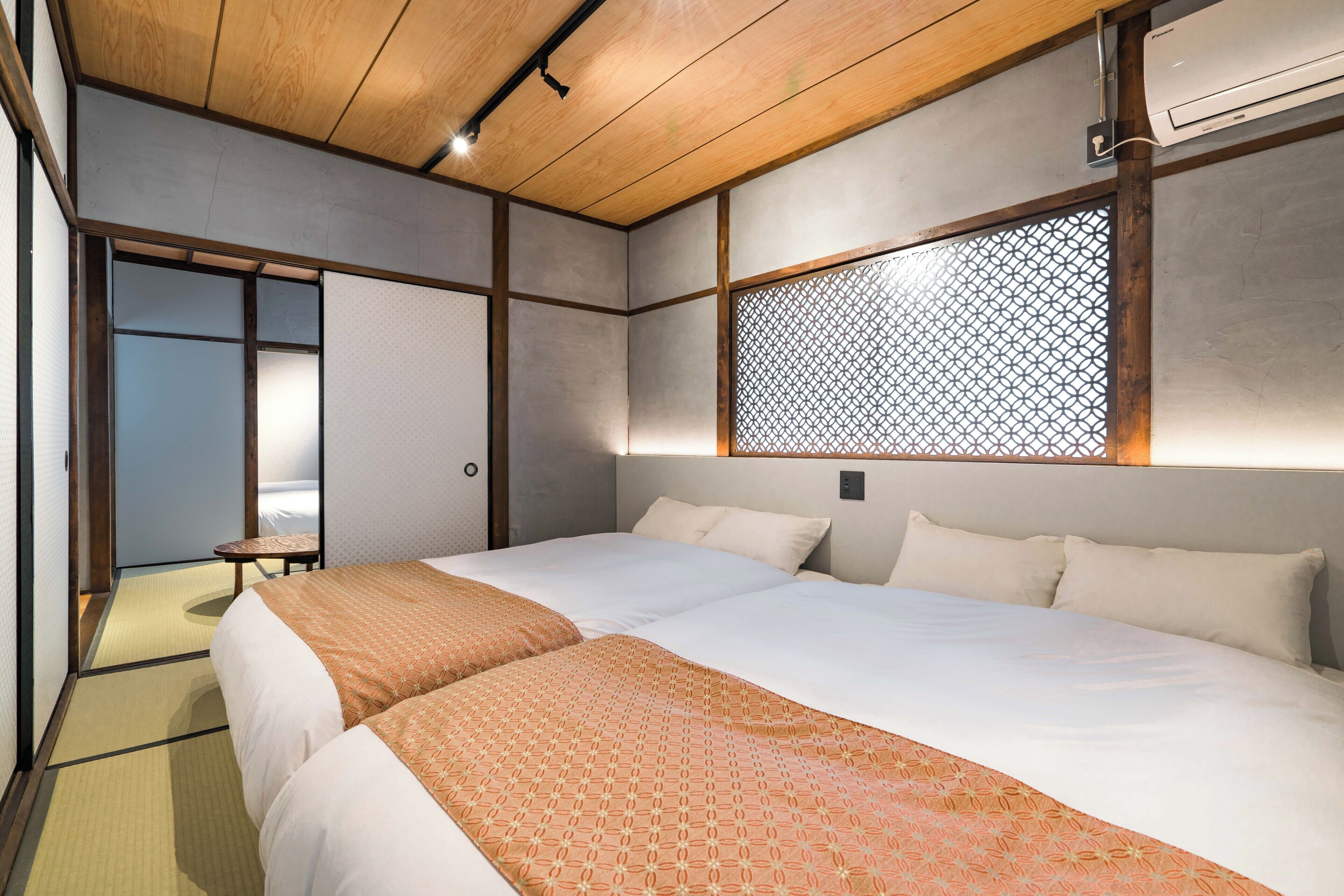 A hotel room with two beds, a wooden ceiling, a wall-mounted air conditioner, and a sliding door leading to a bathroom.