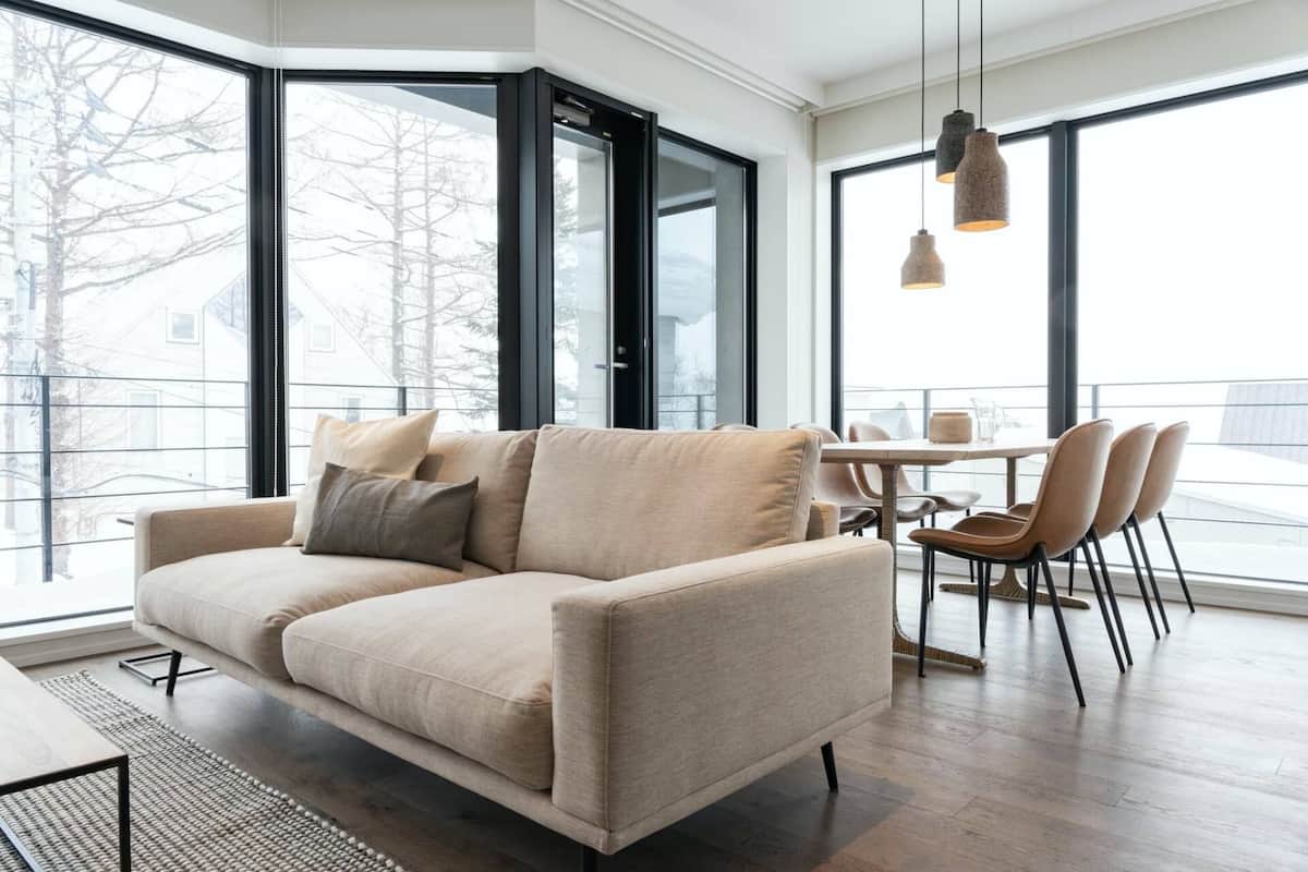 Ein modernes Wohnzimmer mit einem beigefarbenen Sofa, einem Esstisch mit Stühlen und großen Fenstern mit Blick nach draußen.