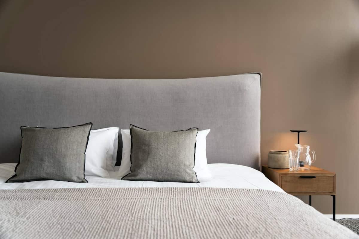 Ein ordentlich gemachtes Bett mit grauem Kopfteil, zwei grauen Kissen und einem weißen Kissen. Ein Holznachttisch mit einer Lampe und einer Vase darauf.