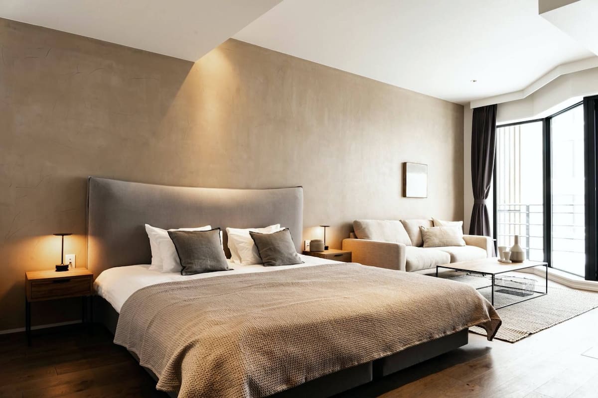 Ein modernes Hotelzimmer mit einem großen Bett, einer kleinen Couch, einem Nachttisch und einem großen Fenster mit Vorhängen.
