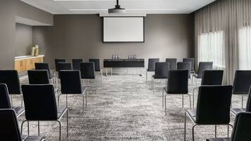 Uma sala de conferência com piso em carpete, cadeiras pretas, uma mesa, tela de projetor e luminária no teto.