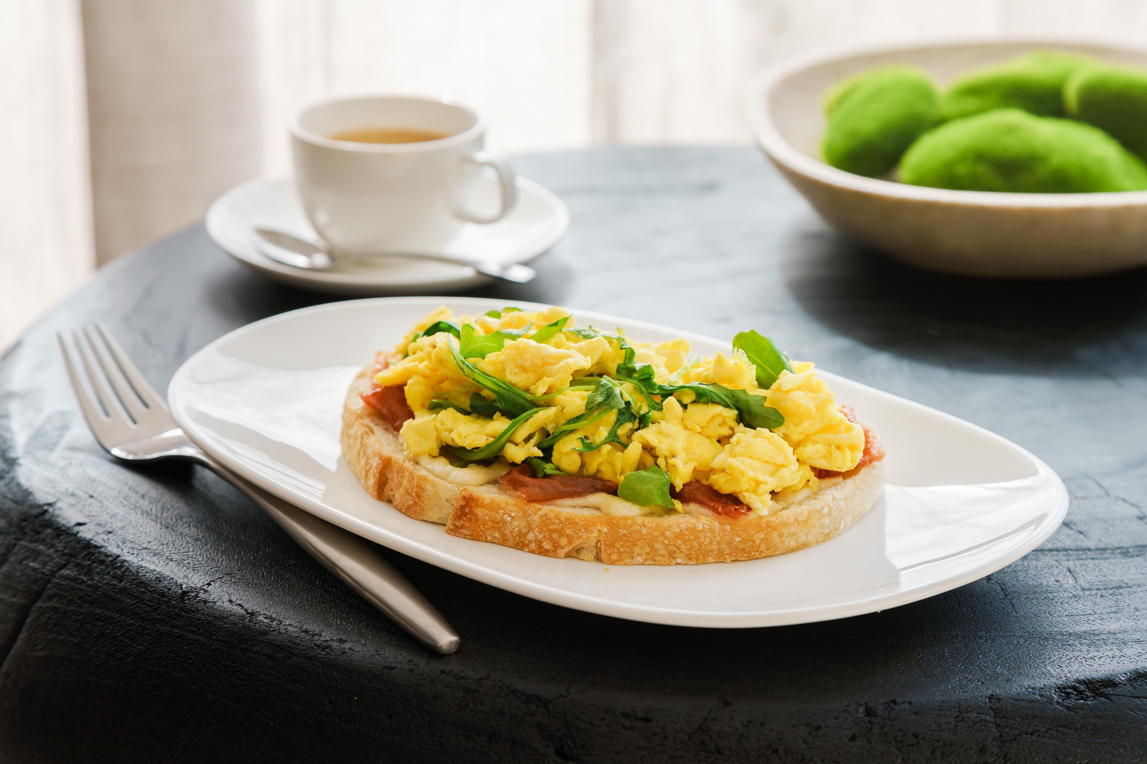 A breakfast plate with scrambled eggs, greens, and ham on toast, a cup of coffee, and a bowl of green dumplings on a dark table.