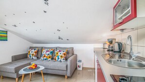 Ein gemütlicher Wohnbereich mit grauem Sofa, bunten Kissen, einem kleinen runden Tisch mit Obst, einer Küchenzeile mit Spüle, Herd und Wasserkocher.