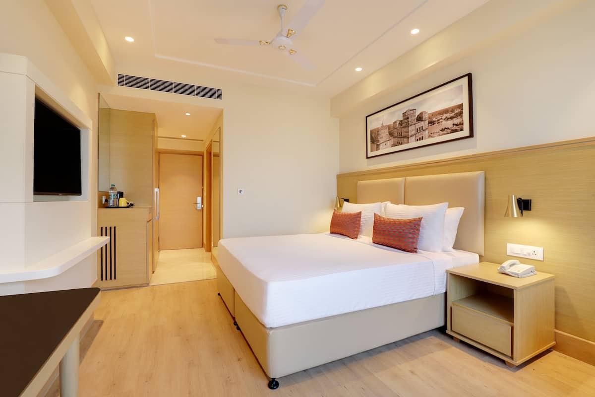 Quarto de hotel com uma cama grande, mesa de cabeceira com abajur, ventilador de teto e um quadro na parede.