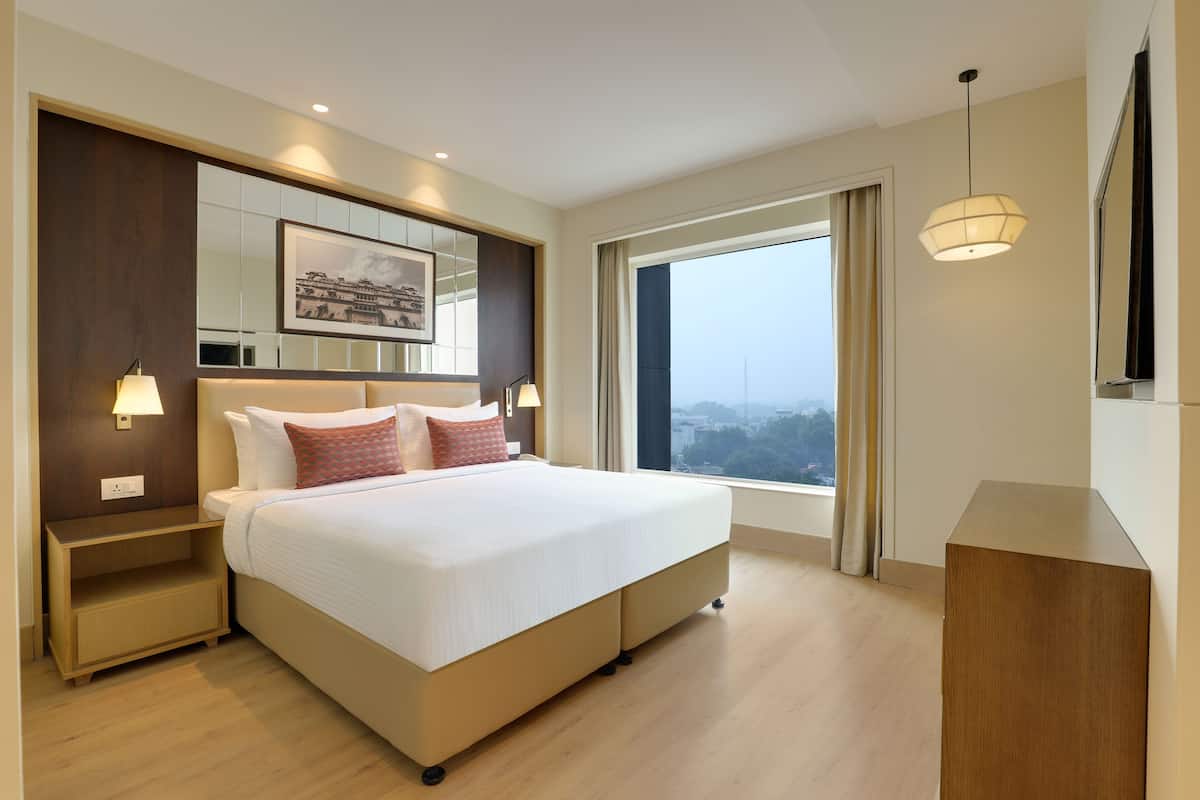 Quarto de hotel moderno com uma cama grande, abajures, uma mesa de cabeceira, uma cômoda de madeira, uma janela ampla com vista e um quadro na parede.
