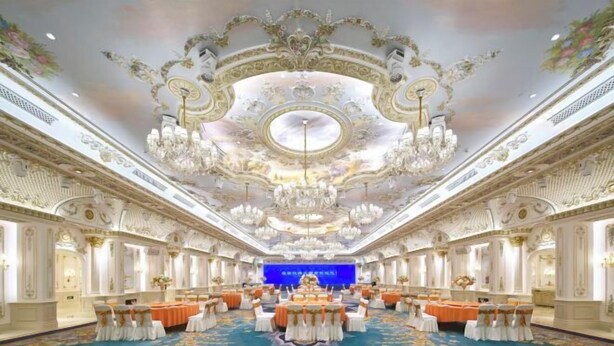 A grand ballroom with ornate ceiling and chandeliers, a central stage, and rows of tables with chairs.