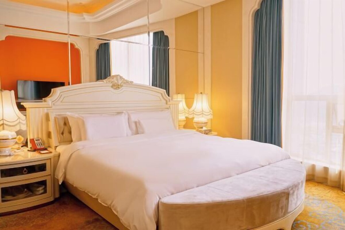 Una habitación de hotel con una cama grande, lámparas de noche, un televisor y un ventanal con cortinas.