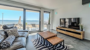A modern living room with a large TV, leather sofa, and a view of the beach.