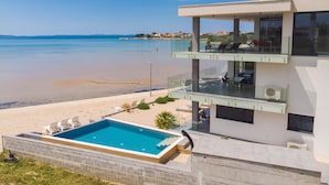 A modern two-story building with a glass balcony overlooking a beach and a swimming pool.