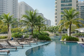 Zona de piscina con sillas de descanso y palmeras, rodeada de edificios altos.