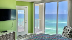 A bedroom with a bed, a TV, a ceiling fan, and a view of the ocean through large windows. - Affordable Beach Front Condo #2 (Panama City Beach)