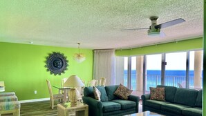 A living room with a green wall, a ceiling fan, a large window with a view of the ocean, and a wicker coffee table. - Affordable Beach Front Condo #2 (Panama City Beach)