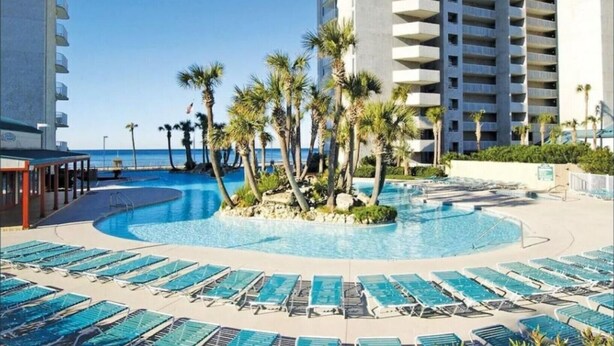 A pool area with lounge chairs and palm trees, surrounded by a high-rise building. - Affordable Beach Front Condo #2 (Panama City Beach)