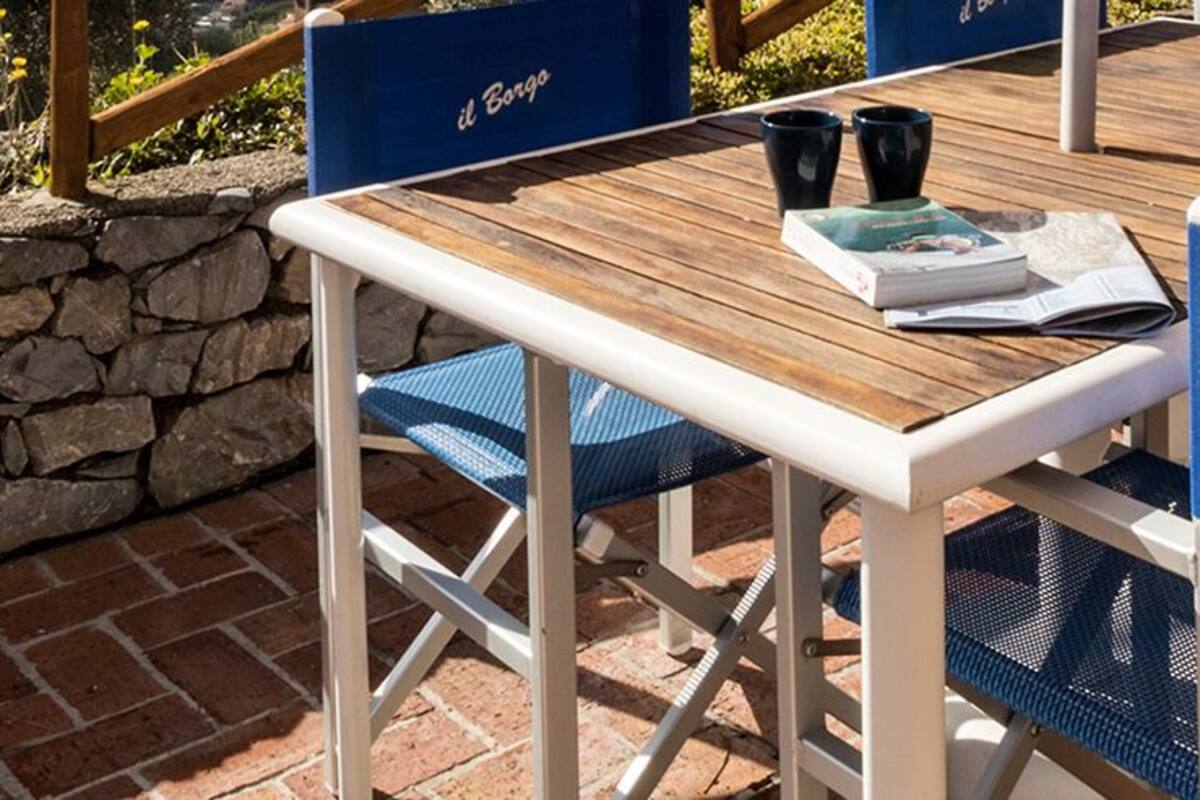 Una terrazza esterna con un muro in pietra, un tavolo in legno e sedie blu.