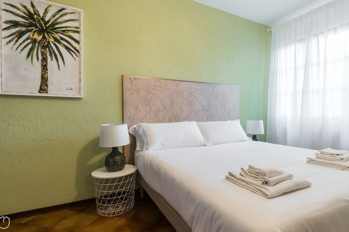 Una camera d'hotel con un letto, due lampade da comodino, un dipinto raffigurante una palma, una testiera a motivi decorativi e una finestra con tende.