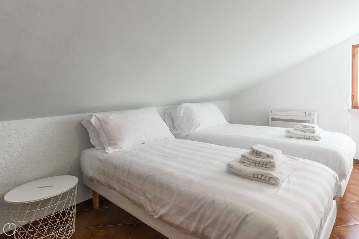 Una camera da letto con due letti, biancheria da letto bianca, un piccolo tavolino rotondo e una finestra con stipiti in legno.