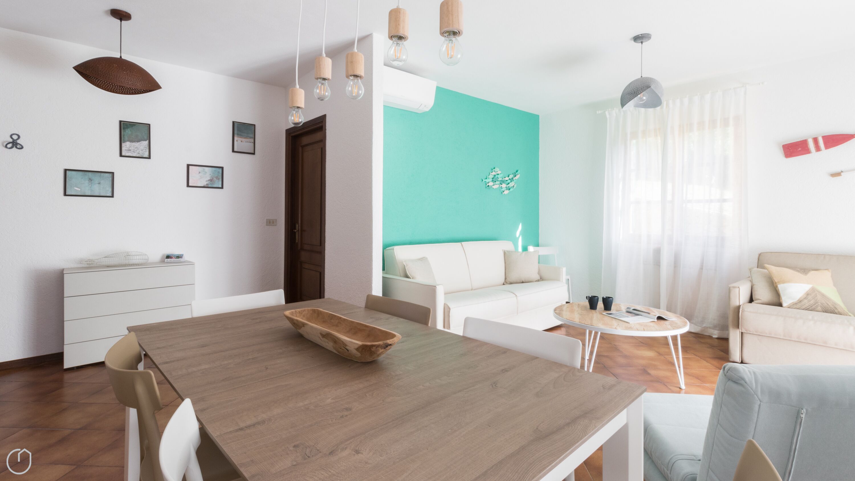 A modern living room with a wooden dining table, white sofas, and a teal accent wall.