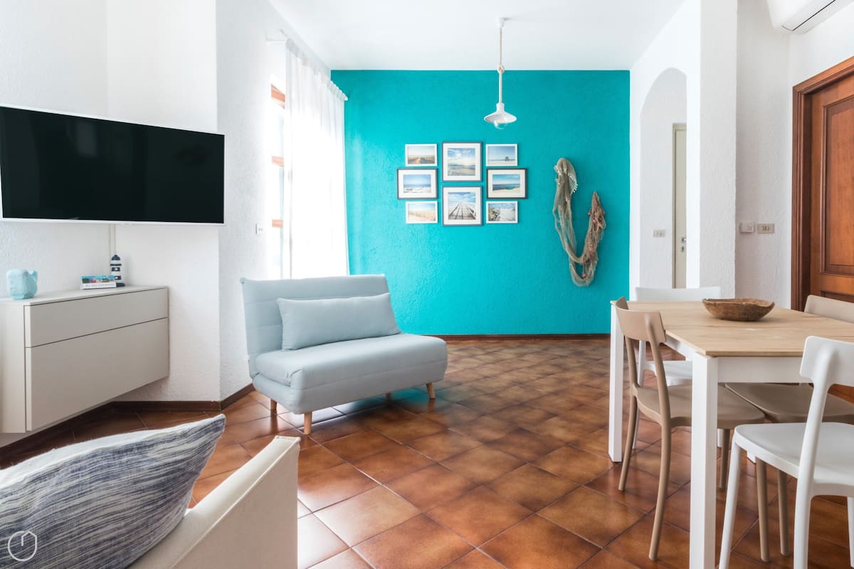 Un soggiorno moderno con una parete verde acqua, una TV a schermo piatto, un divano, un tavolo da pranzo con sedie e una porta in legno.