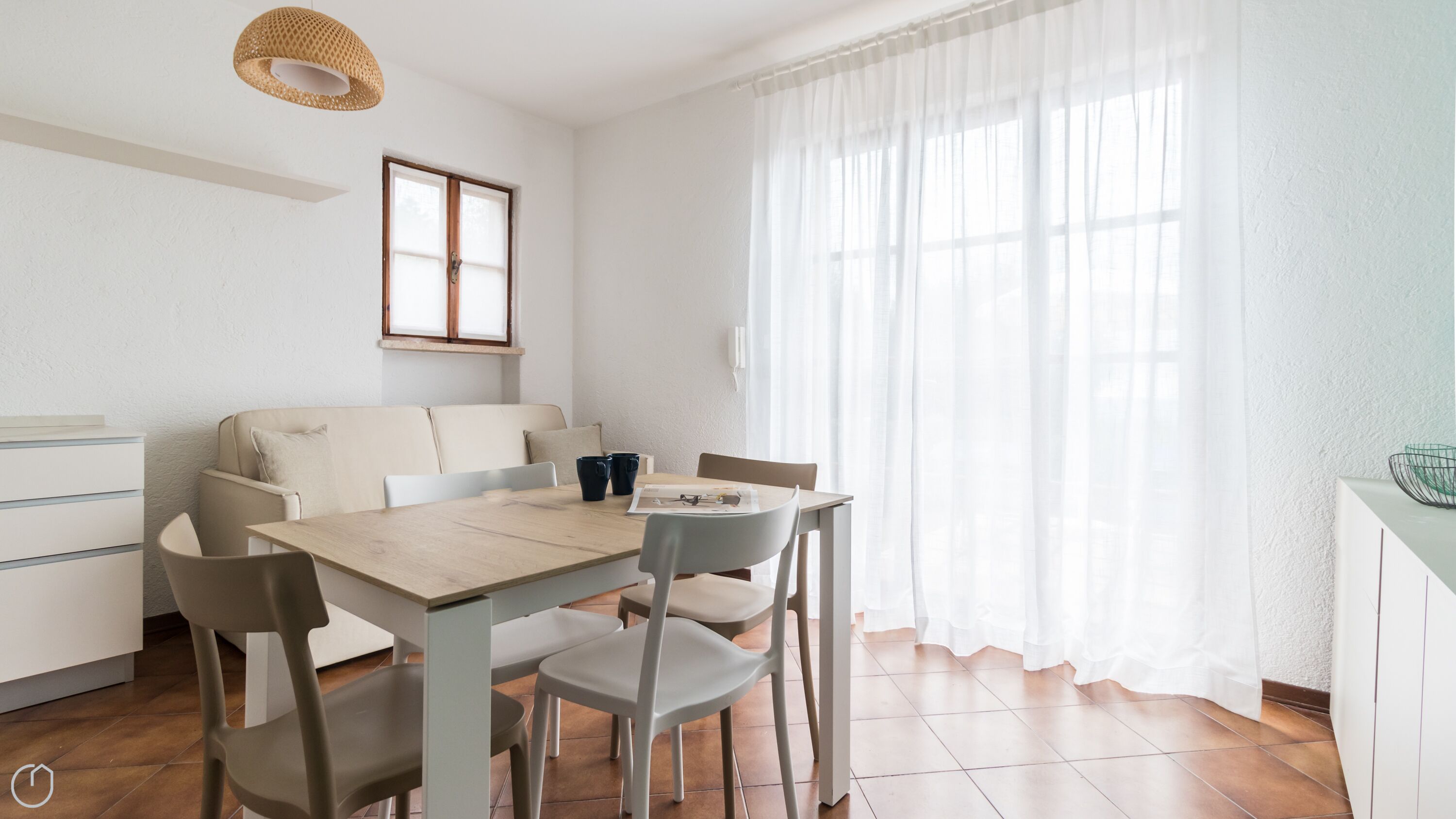 Une pièce moderne et bien éclairée, avec une table à manger, des chaises, un canapé et une fenêtre ornée de rideaux légers.