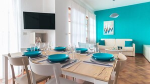 A dining area with a wooden table set for four, surrounded by white chairs. There is a television mounted on the wall and a sofa with a teal cushion in the living area. - Italianway - Il Borgo apartments (Finale Ligure)