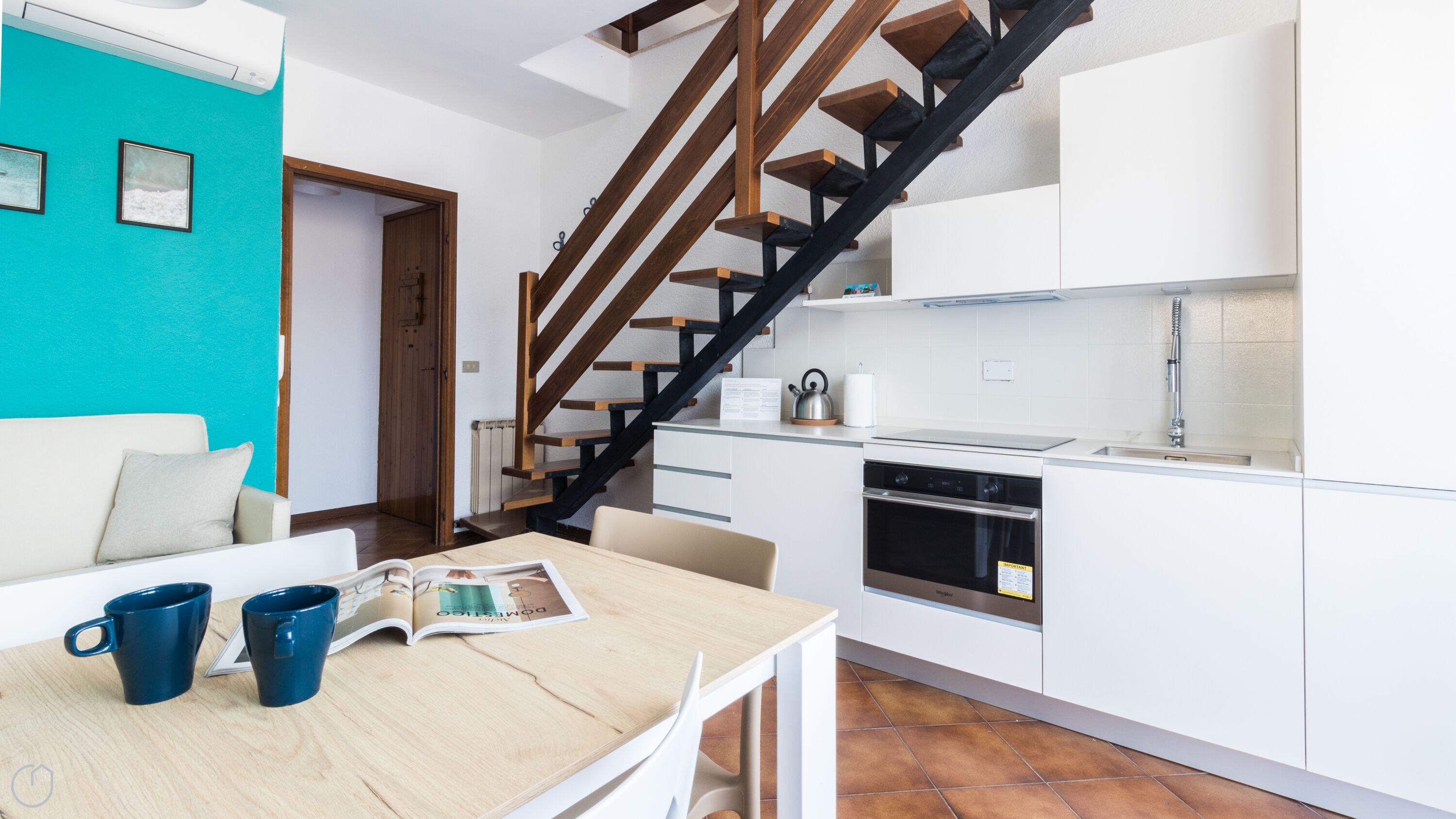 A modern kitchen with white cabinets, a built-in oven, and a stainless steel sink. A wooden staircase with black railings leads to a dining area with a table and chairs.