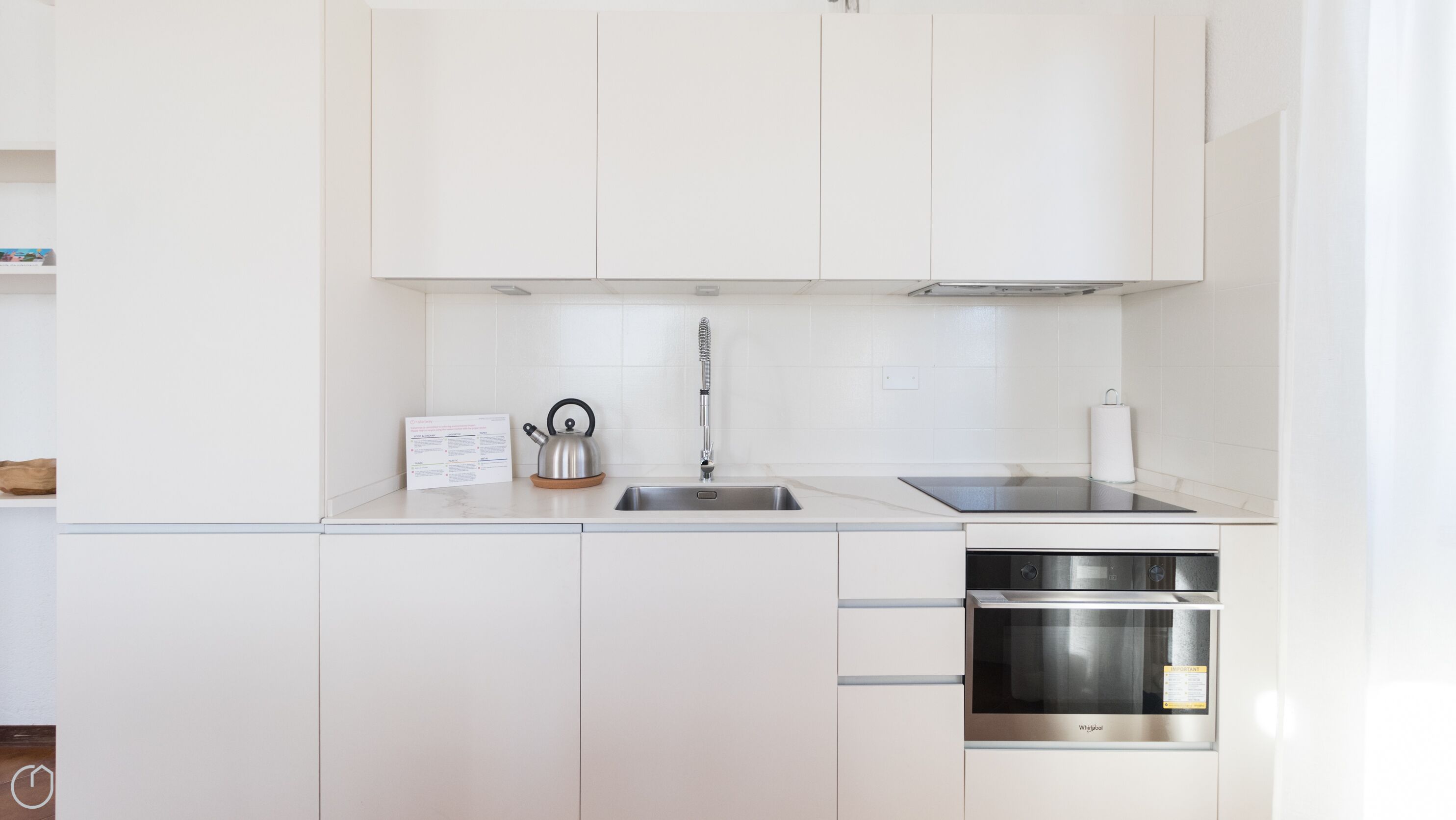 Une cuisine moderne avec des meubles blancs, un évier en acier inoxydable, une bouilloire et un four encastré.