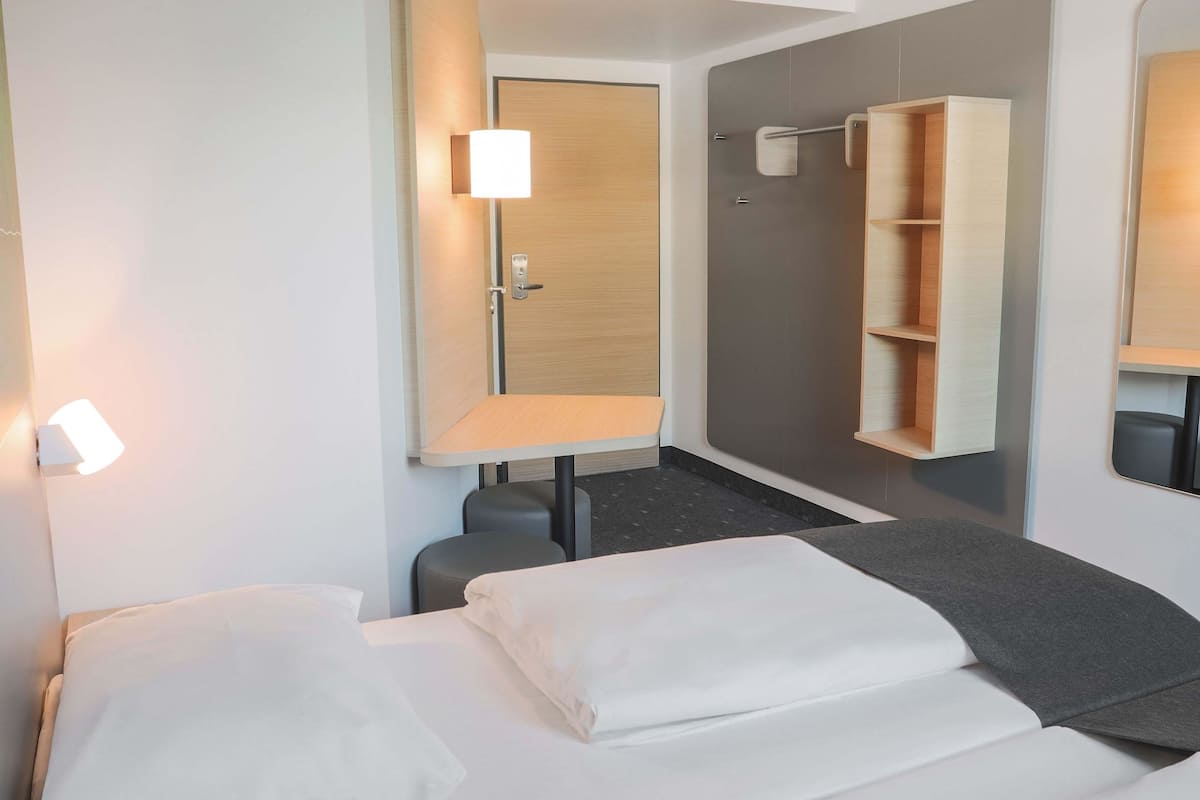 A hotel room with a bed, a desk, a chair, a mirror, and a shelf.