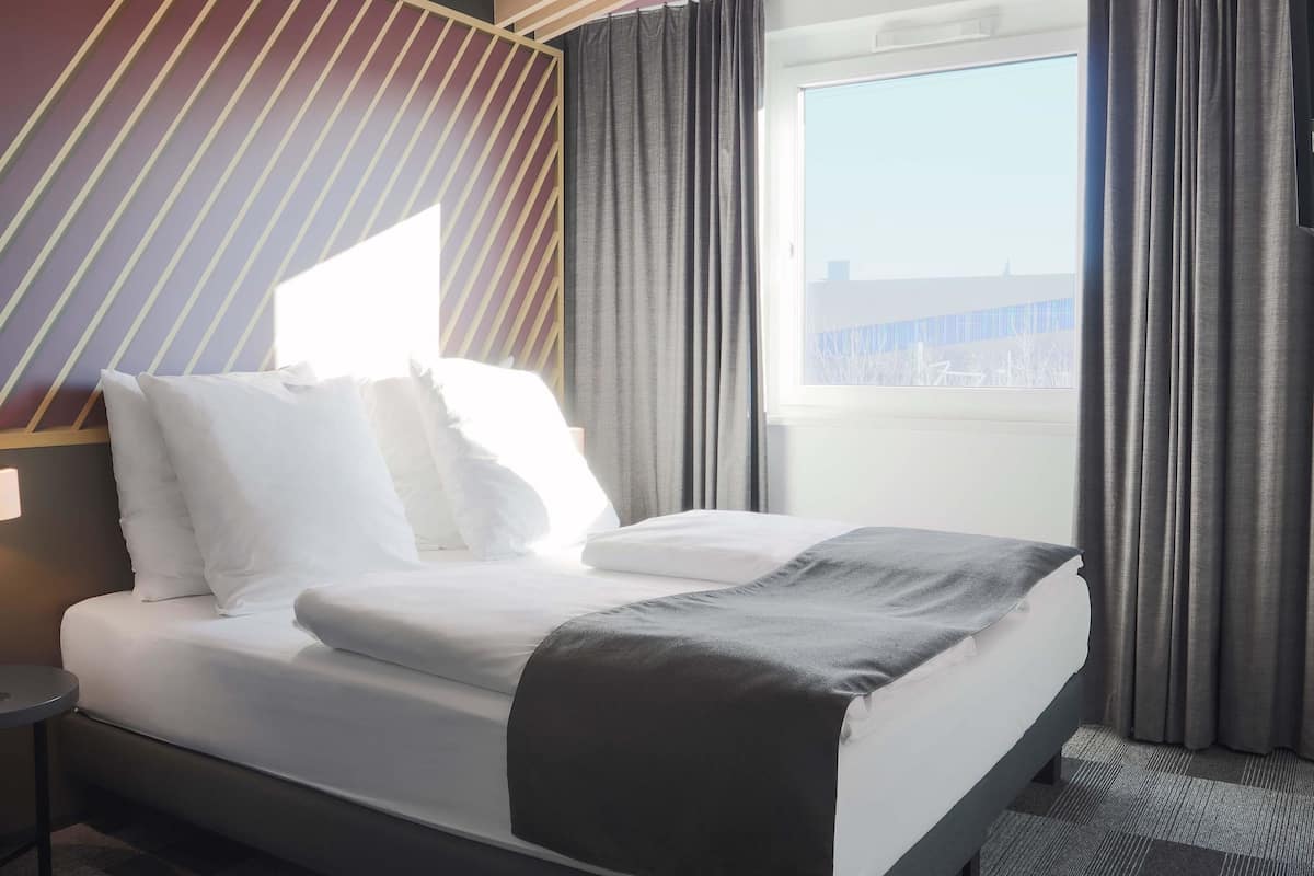 A hotel room with a bed, a nightstand, a window with curtains, and a view of the city.