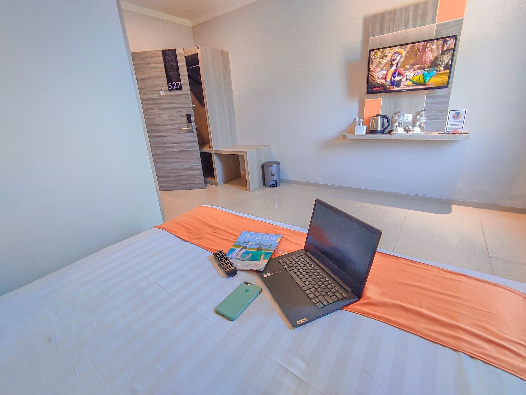 A hotel room with a bed, a laptop, a remote control, and a smartphone on the bed. There is a TV mounted on the wall and a shelf with various items.
