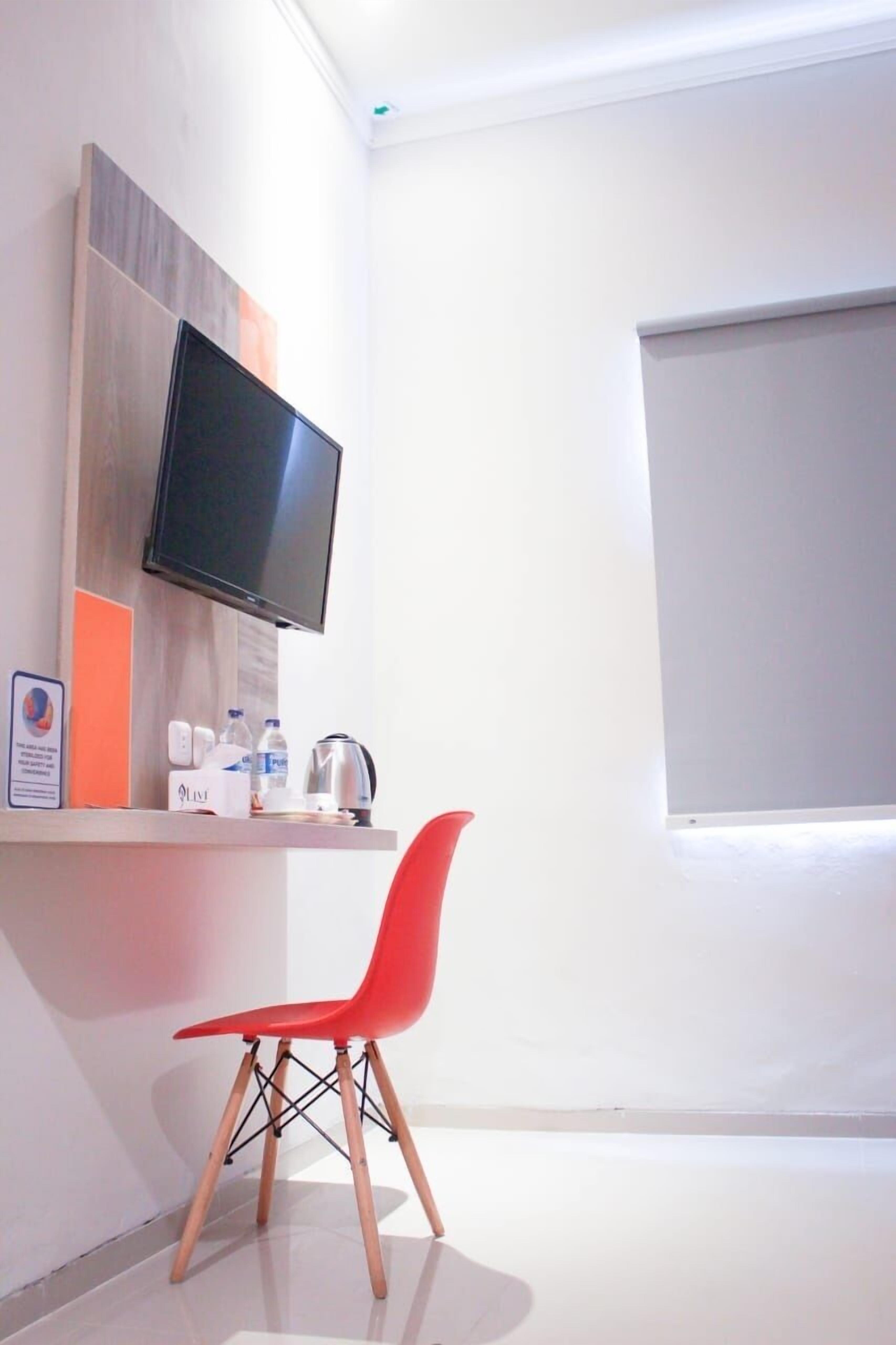 Una habitación moderna con televisión de pantalla plana en la pared, una silla roja, una mesita con una tetera y una ventana con persiana.